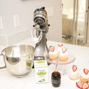 May include: A silver stand mixer with a whisk attachment, a stainless steel mixing bowl, and cupcakes topped with strawberries. Also visible are vanilla extract and a glass cake stand. The scene suggests baking.