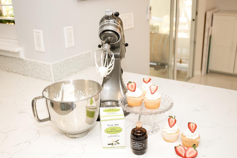 May include: A silver stand mixer with a whisk attachment, a stainless steel mixing bowl, and cupcakes topped with strawberries. Also visible are vanilla extract and a glass cake stand. The scene suggests baking.