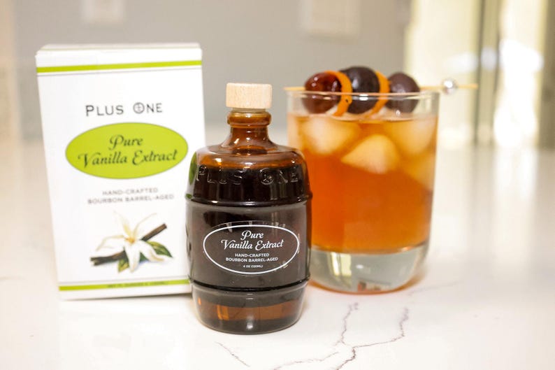 May include: A bottle of Pure Vanilla Extract, with a box and a cocktail. The extract bottle is brown with a wooden cap. The box is white with green text. The cocktail is in a clear glass with ice and a garnish.