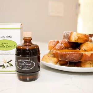 May include: A bottle of pure vanilla extract and a stack of French toast on a white plate. The extract bottle is dark brown with a cork stopper and a label that reads "Pure Vanilla Extract." The French toast is golden brown and drizzled with syrup.