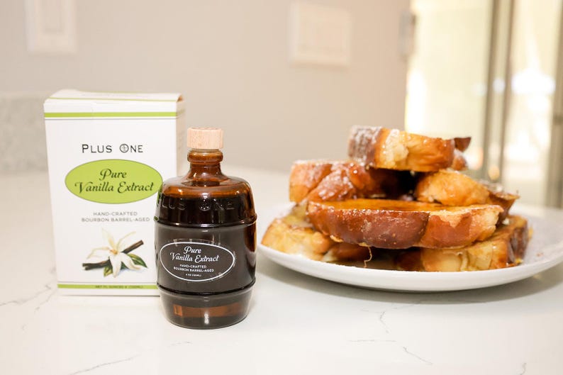 May include: A bottle of pure vanilla extract and a stack of French toast on a white plate. The extract bottle is dark brown with a cork stopper and a label that reads "Pure Vanilla Extract." The French toast is golden brown and drizzled with syrup.