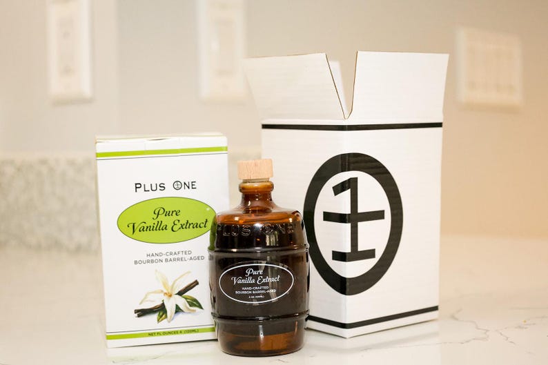 May include: A bottle of Pure Vanilla Extract with a wooden stopper, next to a box with the text "Pure Vanilla Extract". The bottle is dark brown, and the box is white with green accents. The packaging includes the text "Hand-Crafted Bourbon Barrel-Aged".