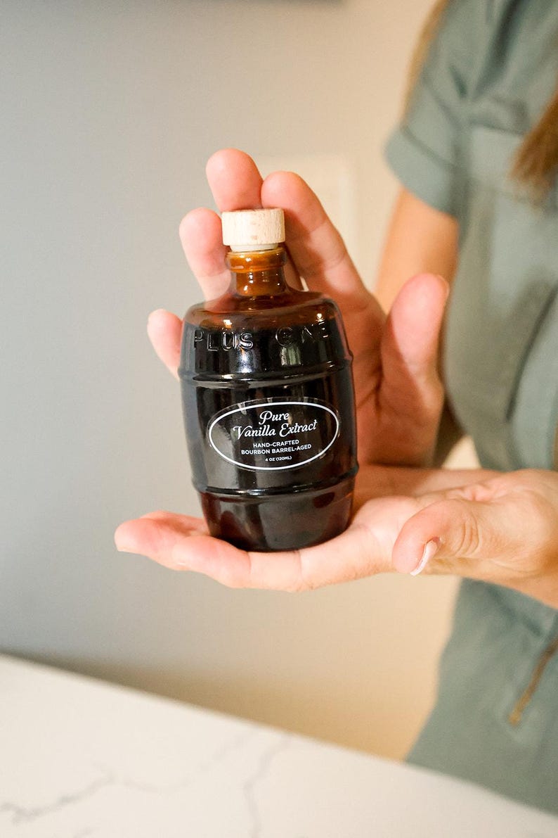 May include: A dark brown, barrel-shaped bottle of Pure Vanilla Extract with a wooden stopper. The label reads "Pure Vanilla Extract, Hand-Crafted, Bourbon Barrel-Aged, 4 fl oz (120ml)." The bottle is held in open hands.