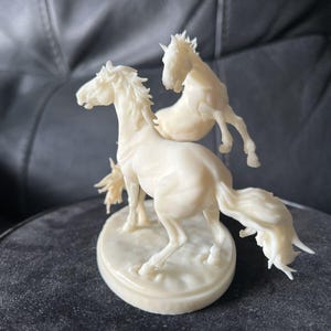 Puede incluir: Una figura de color crema que muestra dos caballos en movimiento sobre una base ovalada. El caballo principal est&aacute; en primer plano con la cabeza girada, mientras que el segundo caballo aparece en una pose encabritada detr&aacute;s.