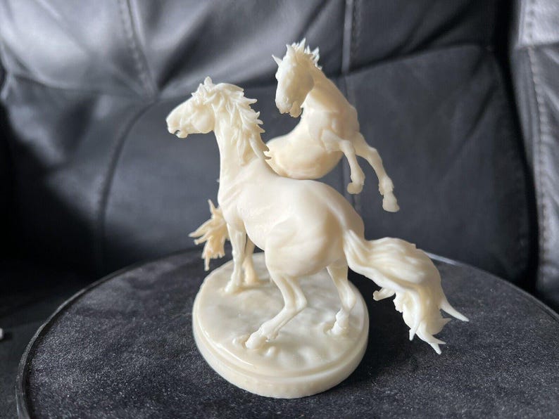 Puede incluir: Una figura de color crema que muestra dos caballos en movimiento sobre una base ovalada. El caballo principal est&aacute; en primer plano con la cabeza girada, mientras que el segundo caballo aparece en una pose encabritada detr&aacute;s.