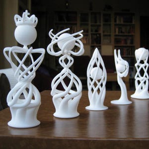 May include: A collection of six white abstract sculptures. Each sculpture has a unique design, featuring spherical elements and intricate, interwoven patterns. The sculptures are displayed on a brown surface, with a blurred background of bookshelves.