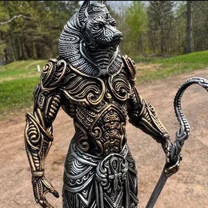 May include: A detailed statue of Anubis, the Egyptian god of the afterlife, with a jackal head and ornate gold and silver armor. The statue holds a staff with a curved end. The figure is standing outdoors with trees in the background.