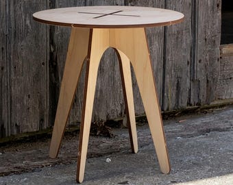 May include: A light-colored wooden side table with a circular top featuring a dark cross-shaped inlay. The table is constructed from layered plywood and stands on three arched legs that meet in the center, creating a stable, open base structure.