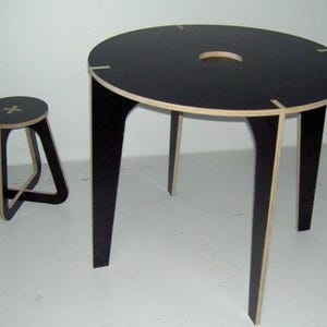 Puede incluir: Un moderno juego de mesa y taburete negros. La mesa redonda tiene una tapa negra con un agujero central y cuatro patas. El taburete tiene un asiento redondo y una base geométrica. Los bordes de la mesa y el taburete son de color claro.