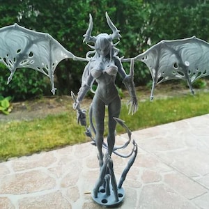 Puede incluir: Figura gris de una demonio femenina con grandes alas, cuernos y un cuerpo detallado. La figura está de pie sobre una base con múltiples zarcillos. Las alas tienen un diseño delicado, similar a una telaraña. La figura está al aire libre.