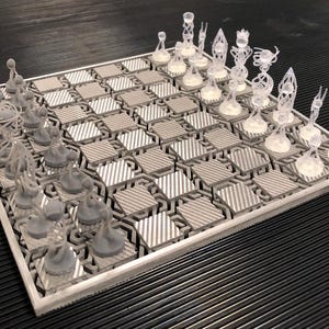 Könnte beinhalten: Ein einzigartiges Schachspiel mit einem geometrischen Brett und durchscheinenden, abstrakten Schachfiguren. Das Brett weist ein silbernes und graues sechseckiges Muster auf. Die Schachfiguren haben komplizierte Designs und verleihen dem klassischen Spiel eine moderne Note.