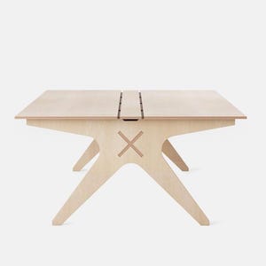 Puede incluir: Una mesa de madera de color claro con una parte superior rectangular y patas anguladas únicas. La mesa presenta un diseño de tapa dividida con un recorte decorativo en forma de X en el lateral. La mesa mide unos 76 cm de largo.