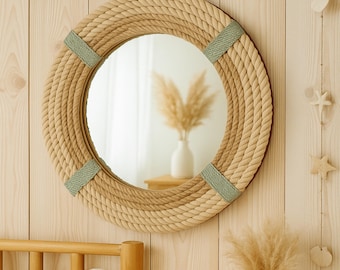 May include: Round mirror with a nautical theme. The mirror is framed with thick, braided rope in shades of beige and tan, accented with light green fabric. The mirror hangs on a light wood wall, creating a coastal aesthetic.
