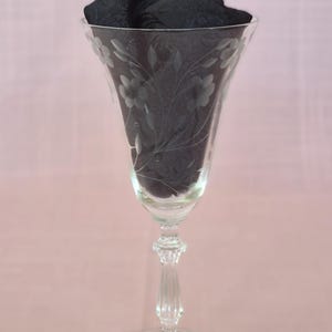 May include: A clear glass goblet featuring an etched floral pattern on the bowl, a multi-faceted stem, and a circular base. The interior of the glass is filled with a dark black fabric, which highlights the delicate etched details on the glass surface.