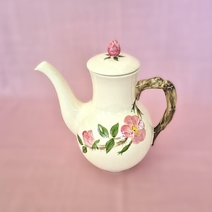 May include: A cream-colored ceramic teapot featuring a hand-painted pink rose design with green leaves on the body. The teapot has a curved spout, a lid topped with a pink rosebud finial, and a handle textured to resemble a brown tree branch.