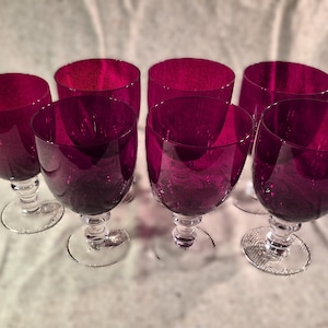 May include: Six stemmed wine glasses with a deep magenta hue. The glasses have a rounded bowl shape and sit on clear, detailed stems and bases. The glasses are arranged on a white surface, creating a visually appealing display.