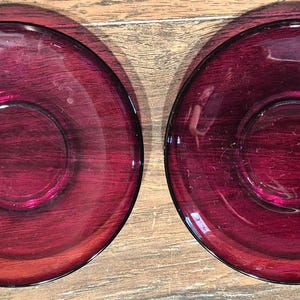 May include: Two matching, deep red glass saucers. Each saucer has a circular indentation in the center and a slightly raised rim. The glass has a glossy finish, reflecting light and creating highlights. The saucers are placed on a wooden surface.