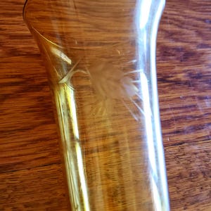 May include: A tall, amber-colored glass vase with a flared top. The vase features a delicate, etched wheat design on its side. The glass has a smooth, polished finish, and the vase is set against a wooden surface.