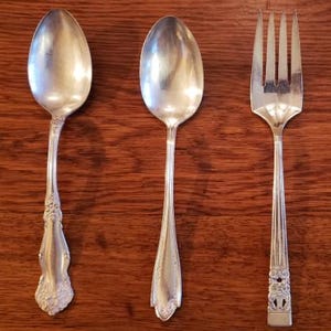 May include: Three pieces of silverware: two spoons and a fork. The spoons have oval bowls and ornate handles. The fork has four tines and a decorative handle. All pieces are silver-colored and rest on a wooden surface.