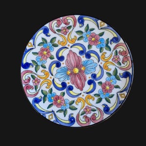 May include: A round, hand-painted ceramic plate with a floral design. The plate features a white background with pink, blue, yellow, and green flowers and decorative swirls. The rim is black.