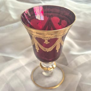 May include: A ruby red wine glass with gold trim. The glass features ornate gold detailing around the rim and on the body. The stem and base are also accented with gold. The glass is likely vintage or antique.