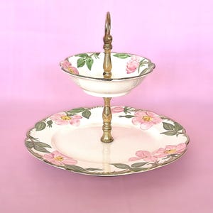 May include: Two-tiered serving dish with a brass handle and support. The white ceramic plates are decorated with pink rose and green leaf patterns. The top tier is a bowl shape, and the bottom tier is a large oval plate.