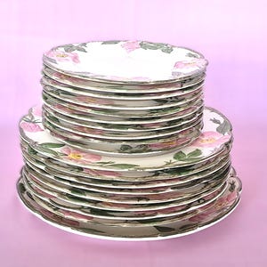 May include: A stack of white ceramic plates featuring a delicate floral pattern of pink rose blossoms and green leaves. The set includes smaller plates stacked on top of larger plates, all with scalloped edges and a thin metallic trim.
