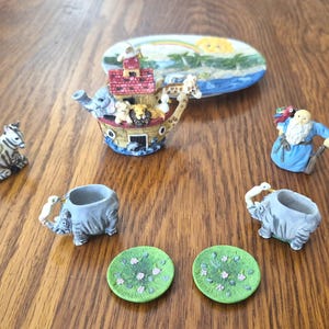 May include: A collection of miniature ceramic figurines including a Noah's Ark boat with animals, a zebra with a blue bird, a bearded figure in a blue robe, two elephant-shaped cups, two green floral saucers, and an oval tray with a rainbow.