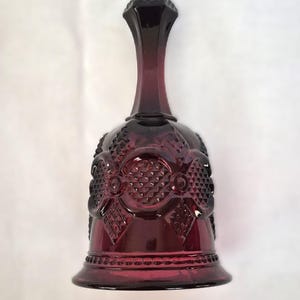 May include: A deep burgundy glass bell featuring a pressed diamond pattern on the body and handle. The bell has a flared rim and a smooth, rounded top section, with circular motifs integrated into the textured geometric glass design.