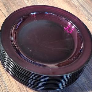 May include: A stack of deep purple glass plates. The plates have a wide rim and a slightly concave center. The color is a rich, dark purple, and the plates appear to be made of a thick glass material.