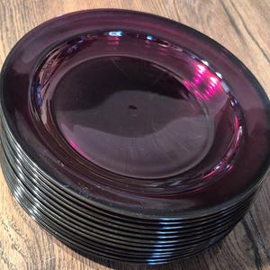 May include: A stack of deep purple glass plates. The plates have a wide rim and a slightly curved shape. The color is a rich, dark purple, and the plates appear to be made of a glossy material. The plates are stacked on top of each other.