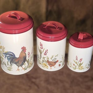 May include: Set of three white metal canisters with red lids and handles. Each canister features a rooster or hen illustration with floral accents. The canisters are cylindrical and vary in size.