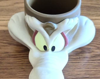 May include: A novelty mug shaped like Wile E. Coyote from Looney Tunes. The mug features a tan-colored cup with a handle, and the coyote's face is painted in cream, yellow, black, and red. The mug is made of ceramic.