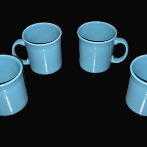 May include: Four light blue ceramic mugs with handles are arranged against a black background. The mugs have a simple, classic design, with a slightly flared rim and a rounded handle. The mugs are evenly spaced.