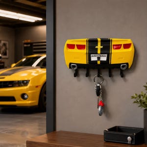 May include: A yellow and black wall-mounted key holder shaped like the rear end of a Chevrolet Camaro. It features red taillights, dual exhaust pipes, and a license plate with the text CAMARO and a small black icon. Five black hooks hold keys.
