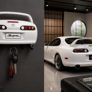Op de afbeelding: Een witte wand-sleutelhouder in de vorm van de achterkant van een Toyota Supra sportwagen. Het is voorzien van de iconische achterspoiler, ronde achterlichten, een Toyota-embleem en een kentekenplaat met de tekst Supra. Zwarte haken houden een autosleutel.
