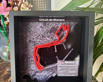 May include: Framed artwork of the Circuit de Monaco. The track layout is highlighted in red against a gray map background. The frame is black, and the text "Circuit de Monaco" is at the top. Additional text provides race details.