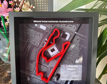 May include: Framed artwork featuring the Miami International Autodrome race track in red, set against a detailed map background. The frame is black, and the artwork includes text indicating the track's location in Florida.