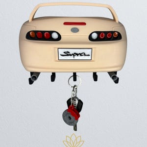 May include: A beige Supra car-shaped key holder with black key hooks and a rear spoiler. The key holder has red and white taillights and the word "Supra" on the license plate. A set of keys with a red brake disc charm hangs from the key holder.
