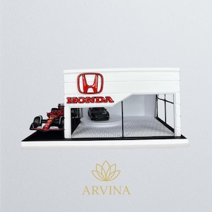 May include: A miniature Honda dealership model with a red Formula 1 race car and a black car inside. The white building features the Honda logo and text in red. The model sits on a black base.