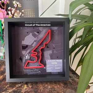Peut inclure: Une œuvre encadrée représentant le circuit des Amériques à Austin, Texas. Le tracé rouge se détache sur un fond de carte gris. Le cadre noir entoure l'œuvre, avec du texte détaillant les spécifications de la piste.