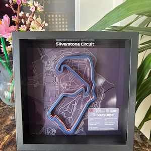 Könnte beinhalten: Ein gerahmter 3D-Plan der Silverstone-Rennstrecke, Großbritannien, mit blau-rotem Streckendesign. Der schwarze Rahmen umgibt die Karte, die Text mit Details zum Grand Prix enthält. Der Text "Silverstone Circuit" steht oben.