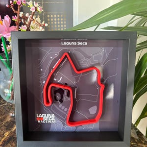 May include: A framed artwork featuring the Laguna Seca Raceway track layout. The track is rendered in red, set against a gray map background. The frame is black, and the text "Laguna Seca Raceway" is visible.
