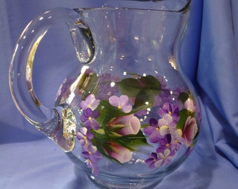 May include: A clear glass pitcher with a large handle and a pouring spout. The pitcher is decorated with hand-painted purple flowers and green leaves. The pitcher is likely used for serving beverages.