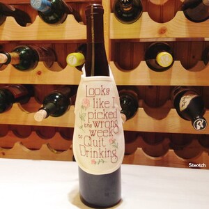 May include: A wine bottle with a decorative apron that reads "Looks like I picked the wrong week to Quit Drinking." The apron is cream-colored with embroidered text and floral accents. The background features a wooden wine rack filled with bottles.