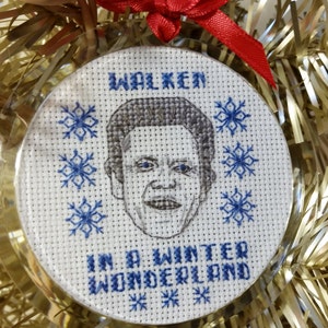 May include: A white round Christmas ornament with a cross-stitch design featuring a portrait of actor Christopher Walken and the text "Walken in a winter wonderland".