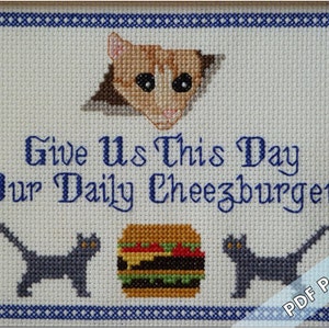 May include: Cross-stitch pattern featuring a cat peeking out of a hole, two cats on either side of a cheeseburger, and the text "Give Us This Day Our Daily Cheeseburger".