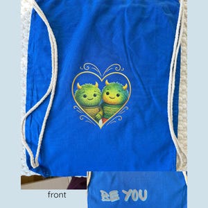 Könnte beinhalten: Blauer Kordelzugbeutel mit einem herzförmigen Design mit zwei grünen Monsterfiguren. Auf der Rückseite der Tasche steht der Text "BE YOU". Die Tasche hat weiße Seilriemen.