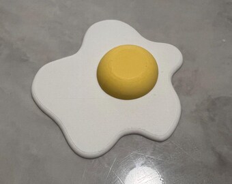 Può includere: Un sottobicchiere bianco a forma di uovo con un cerchio giallo al centro, che ricorda un uovo fritto. Il sottobicchiere è realizzato in un materiale solido e poggia su una superficie grigia. Il design è semplice e giocoso.