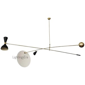 Op de afbeelding: Een moderne kroonluchter uit het midden van de eeuw met een messing afwerking. De armatuur heeft een zwart en goud kegelvormig licht, een witte schijf en een gouden bol. De armen strekken zich horizontaal uit en creëren een evenwichtig, artistiek ontwerp.